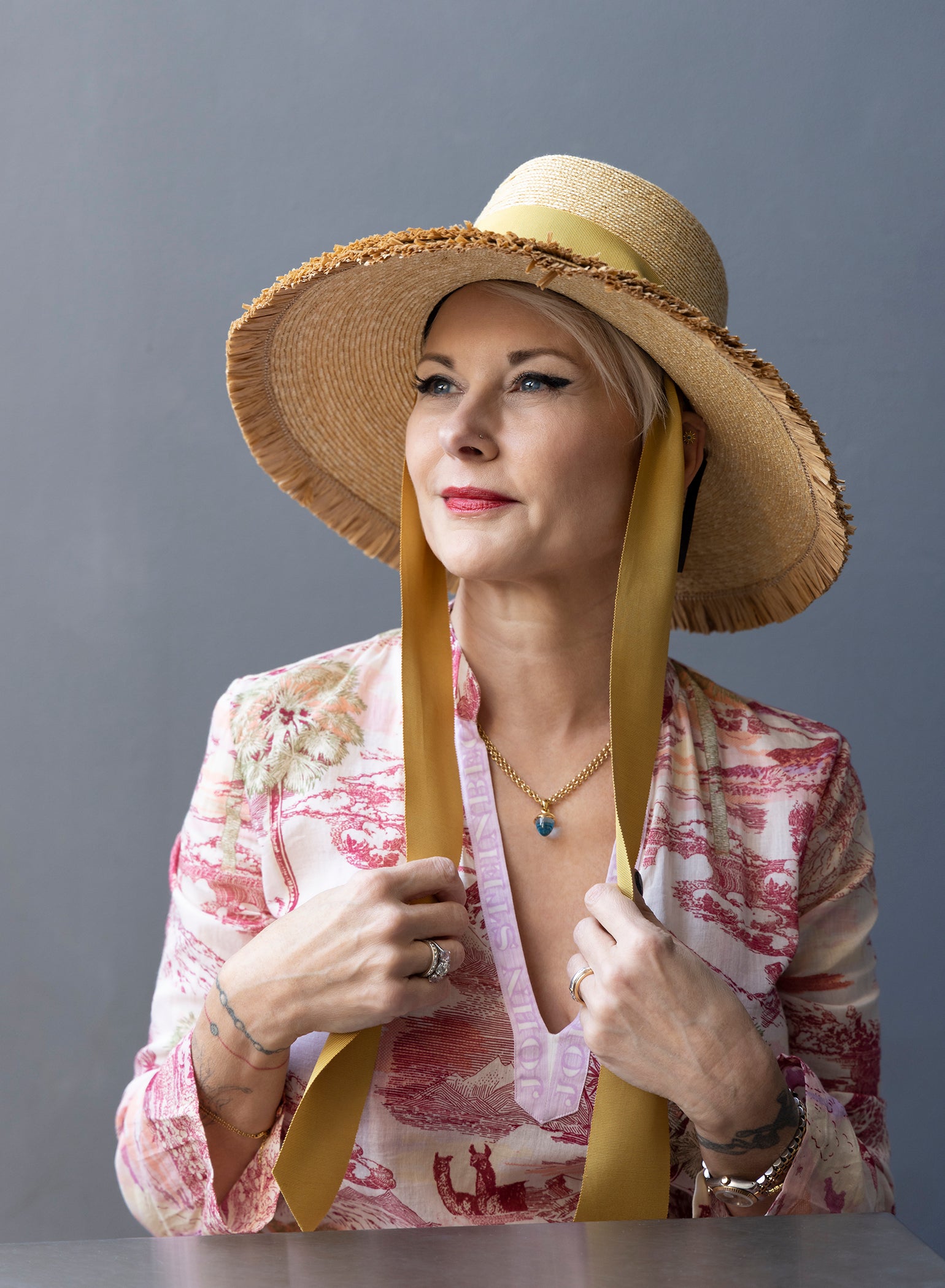 Straw beach hat with double row of fringe on the brim. Crown has two gold grommets on the sides with a matching wide ribbon threaded through the grommets. Hat is shown on a model holding the ribbon.