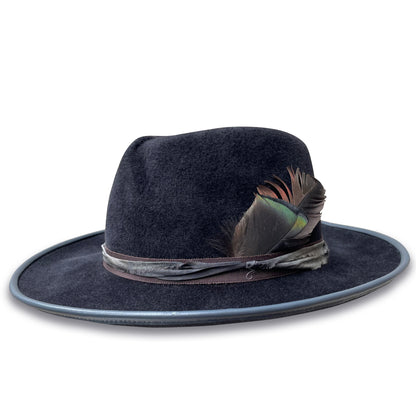 Charcoal fur felt fedora trimmed with brown ribbon layered with silk sari fabric. Finished with  turkey feathers. Brim is piped with matching leather. Crown is pleated and brim is 2.75" wide. 